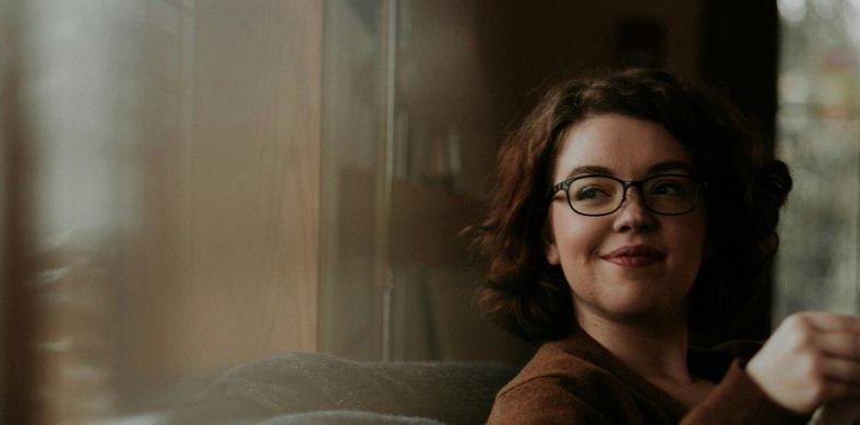 A woman with glasses and curly hair gently smiles while holding a book in a warm and cozy setting.
